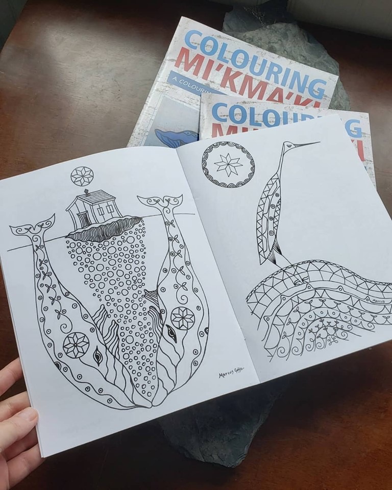 A book is held open showing black line art of two whales with a house between them on one page, and a crane on the opposite. Two copies of "Colouring Mi’kma’ki" are stacked in the background.