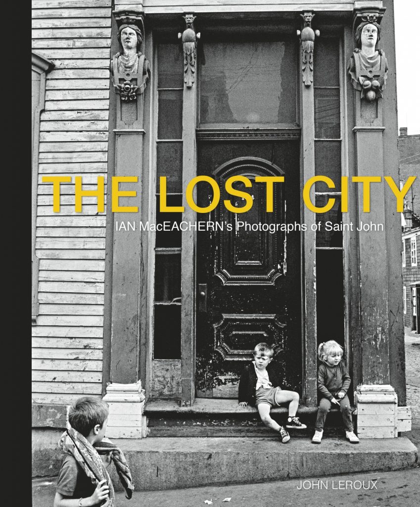 A black and white photograph of two children sitting on the front step of a building , with a third child looking on. The Lost City is printed across the centre in yellow all-caps with the subtitle in white printed below: "Ian MacEachern's Photographs of Saint John". The author's name, John Leroux, is printed in white all caps in the bottom right corner.