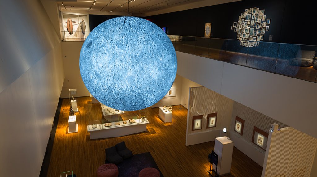 A glowing blue moon hangs across from a second-story balcony in a darkened gallery. Various art works are visible both on this second story and below in the first.