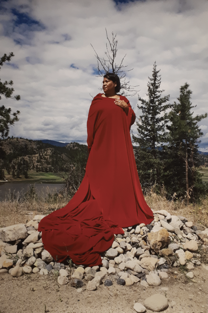 Lori Blondeau, regal, stands dressed in a long red cloth wrapped into dress or robe, on a pile of rocks, in a landscape of trees, hills, and water. The artist looks to the side, left hand on chest.